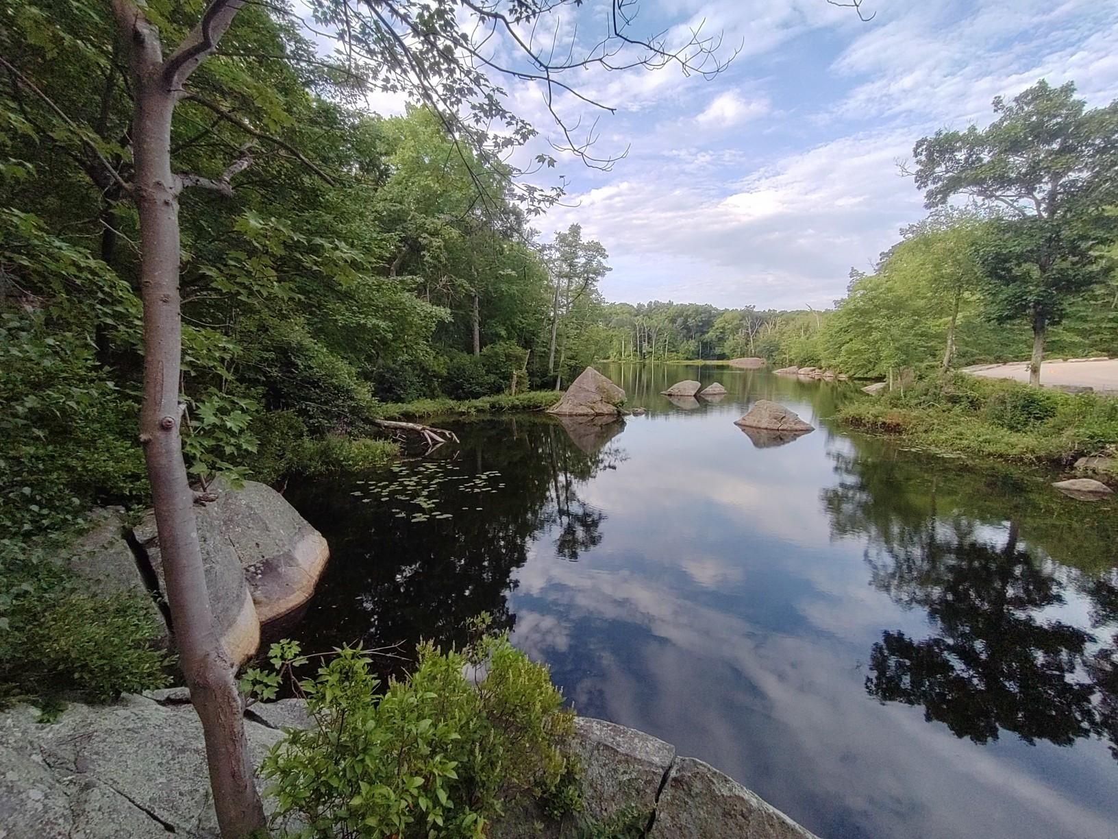 Calm lake with large rocks and green trees under a cloudy sky. - Pond Drop Storm Water Pond loop from Lincoln Woods State Park