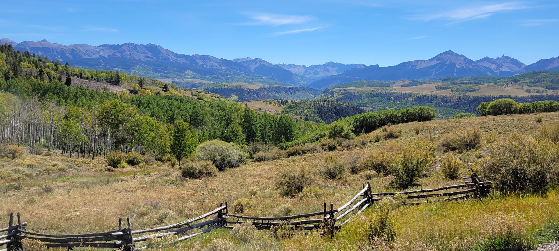 Sunny mountain valley with green and golden trees, distant peaks, and a wooden fence.