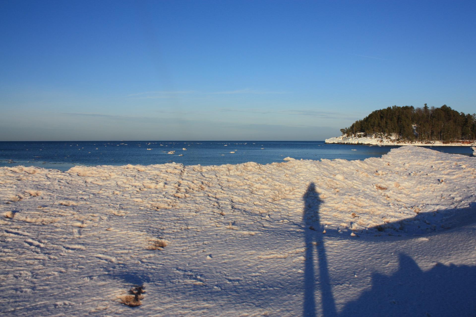 A long human shadow stretches across a snowy beach next to blue water.
