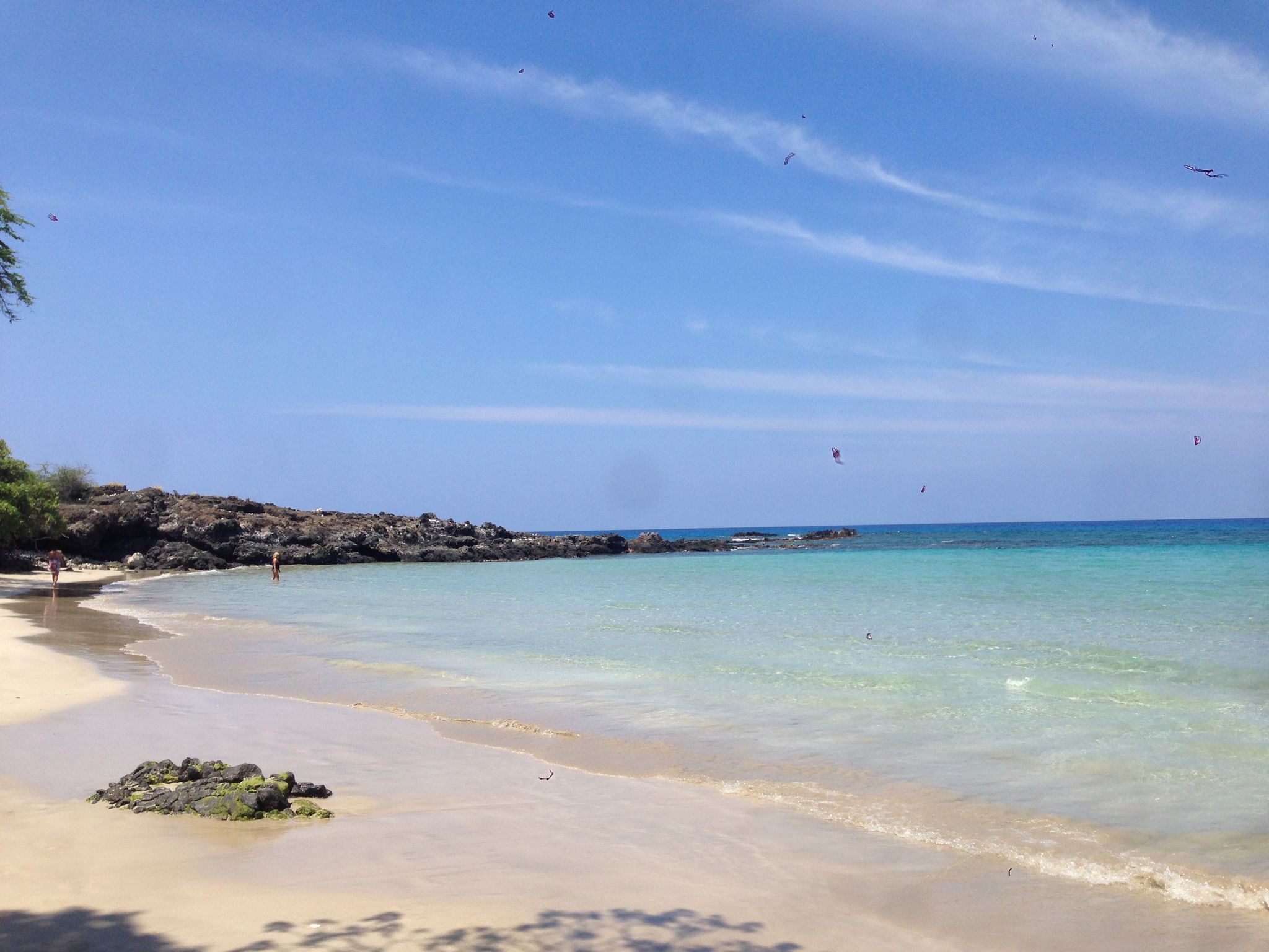 A sunny beach with clear turquoise water, rocky shore, and distant people. - Lulahala Point to Waimea loop