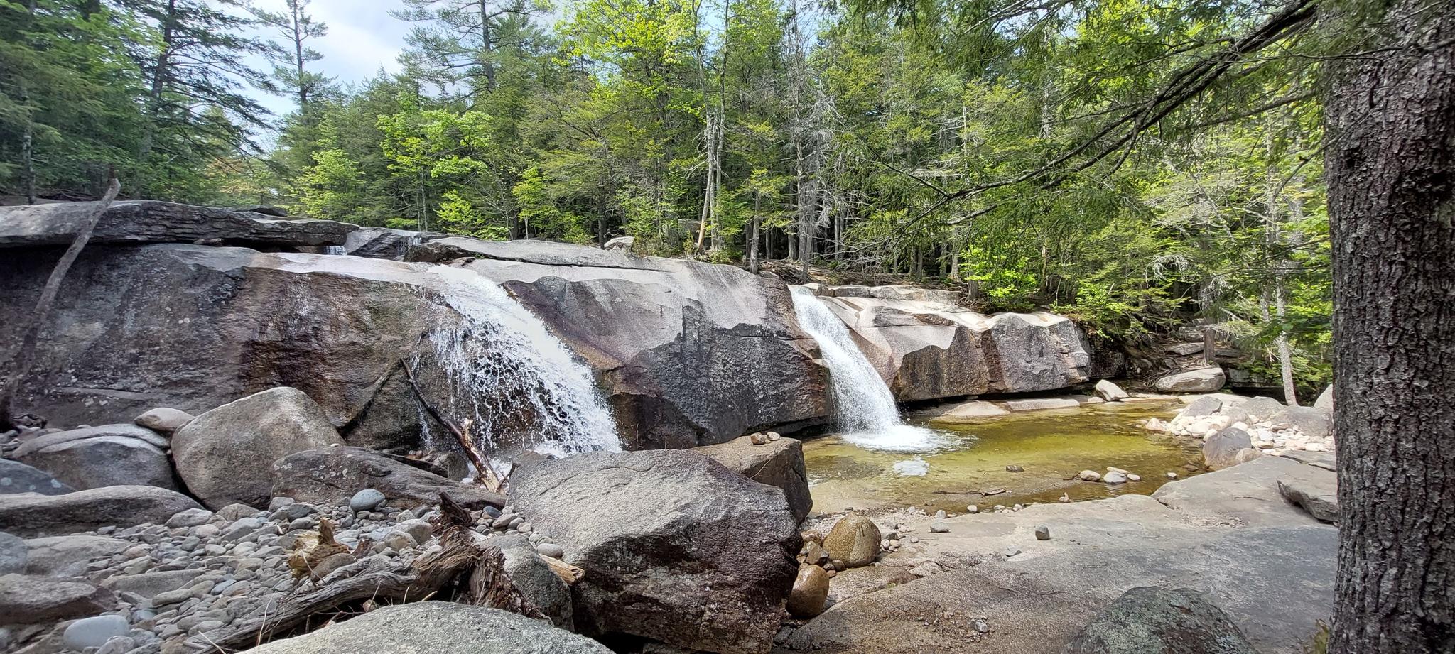Two waterfalls cascade over large, flat rocks in a lush green forest.