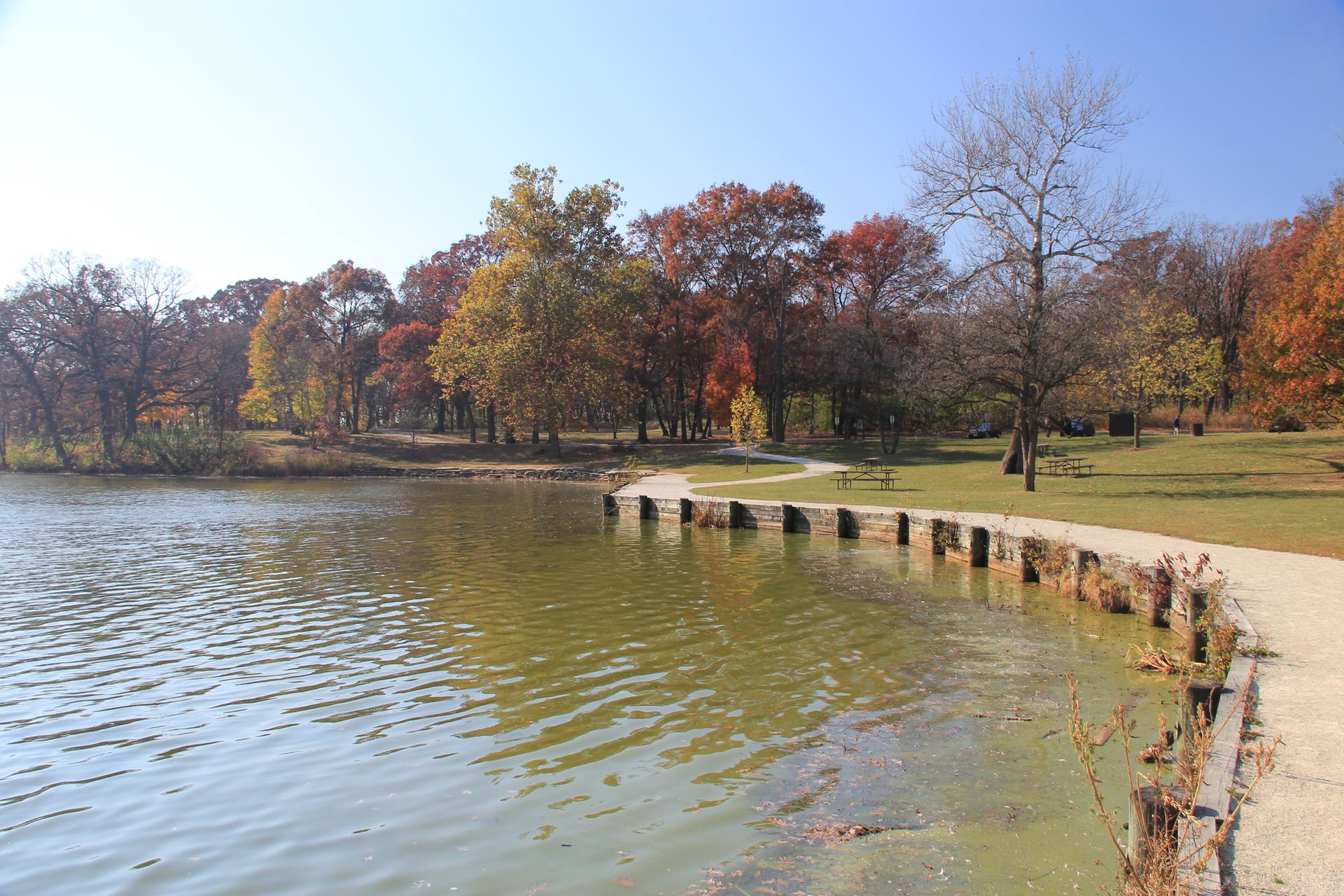 A lake with a paved path, surrounded by colorful autumn trees on a clear day.