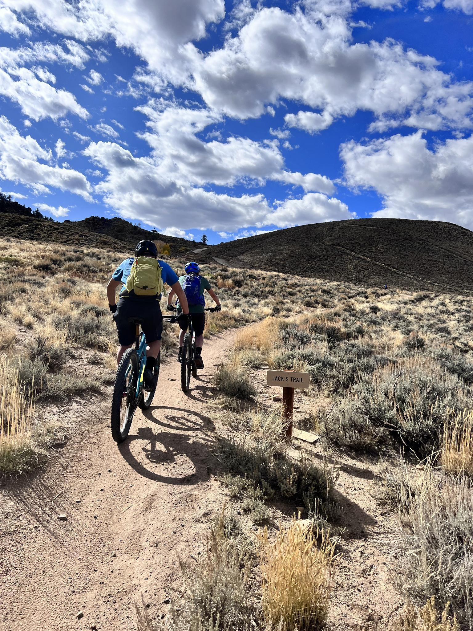 Two people mountain biking on a dirt trail under a bright sky.