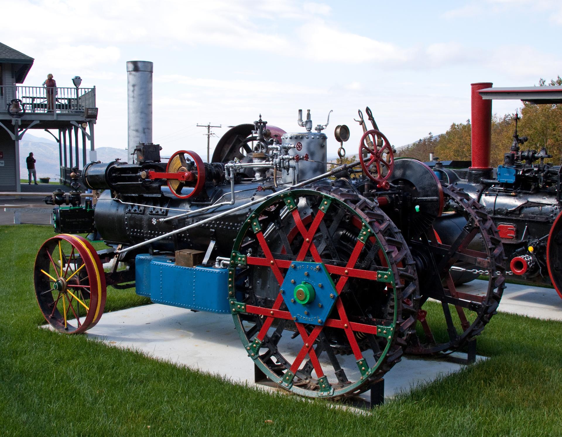 A large, colorful vintage steam tractor on grass under a cloudy sky. Two people visible.
