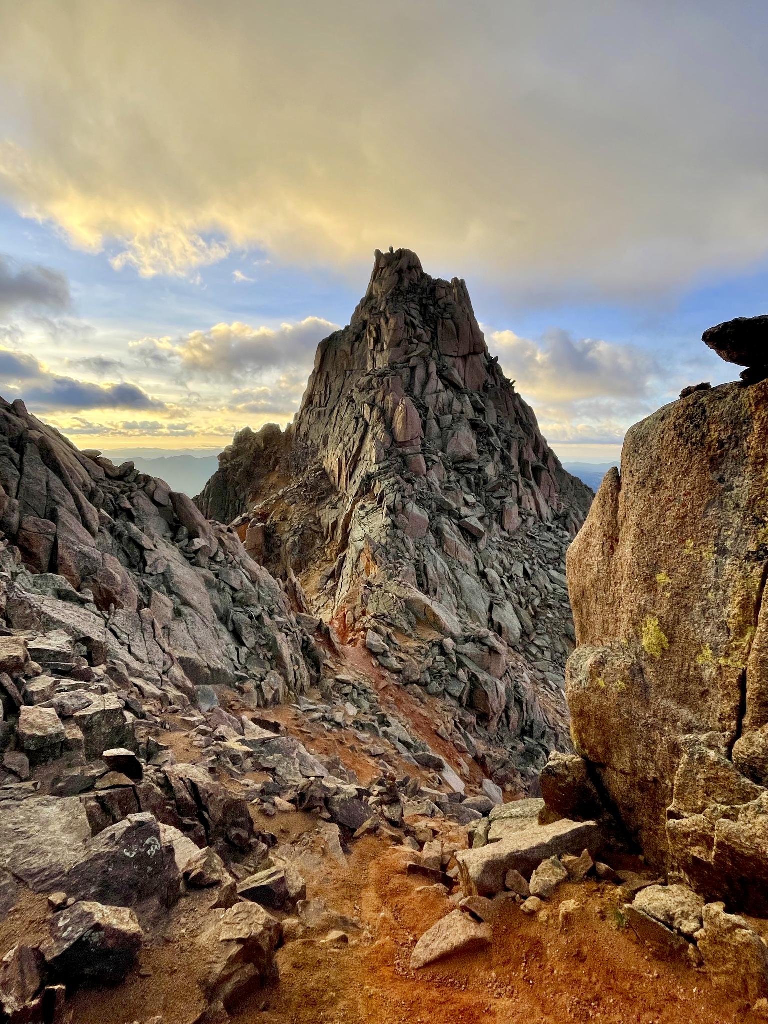 Dramatic rocky peak rises from a steep, rugged landscape under a cloudy sky.
