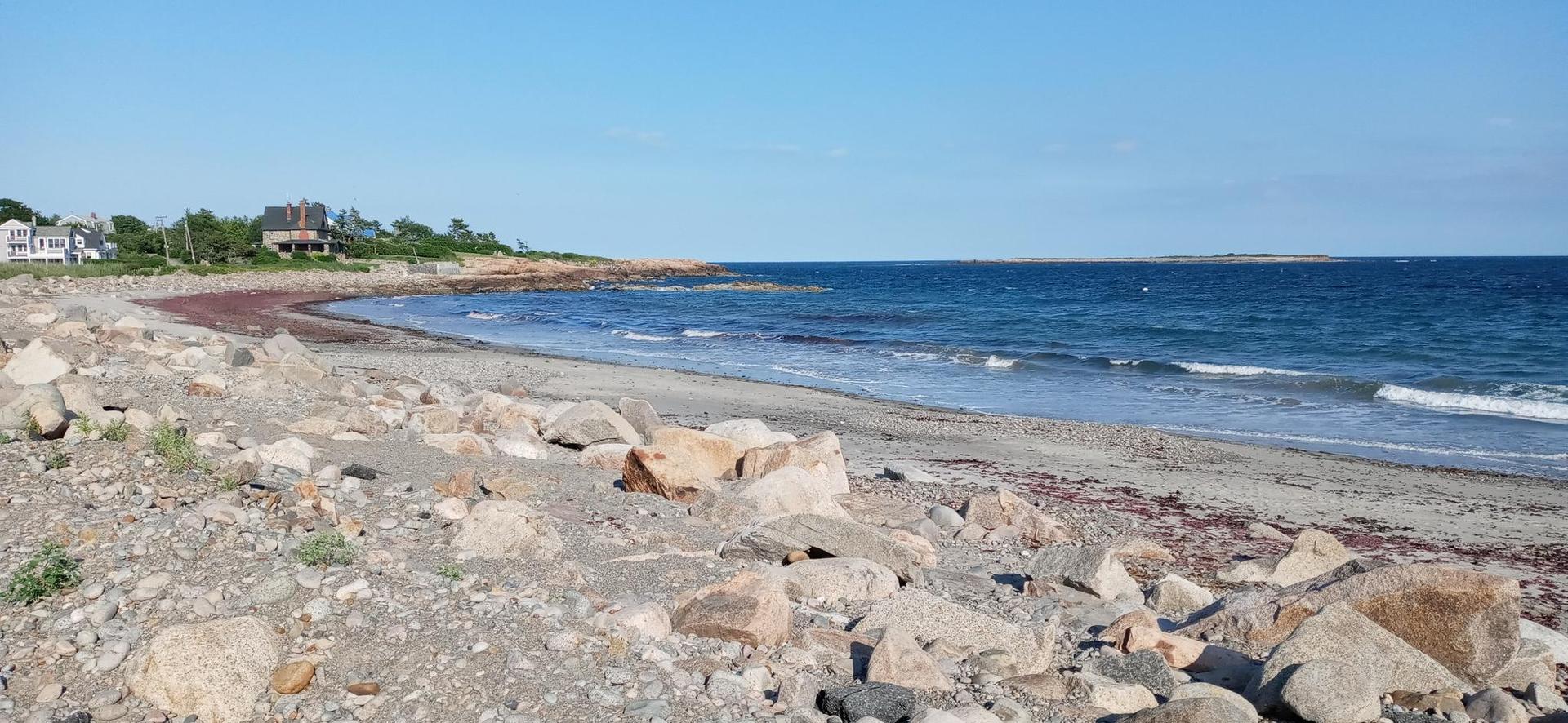 Rocky beach with gentle waves, distant houses, and a clear blue sky.