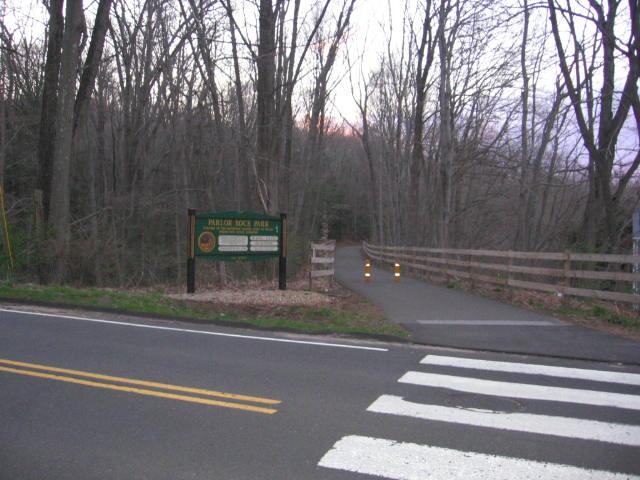 A paved road and crosswalk lead to a wooded path with a sign and fence.