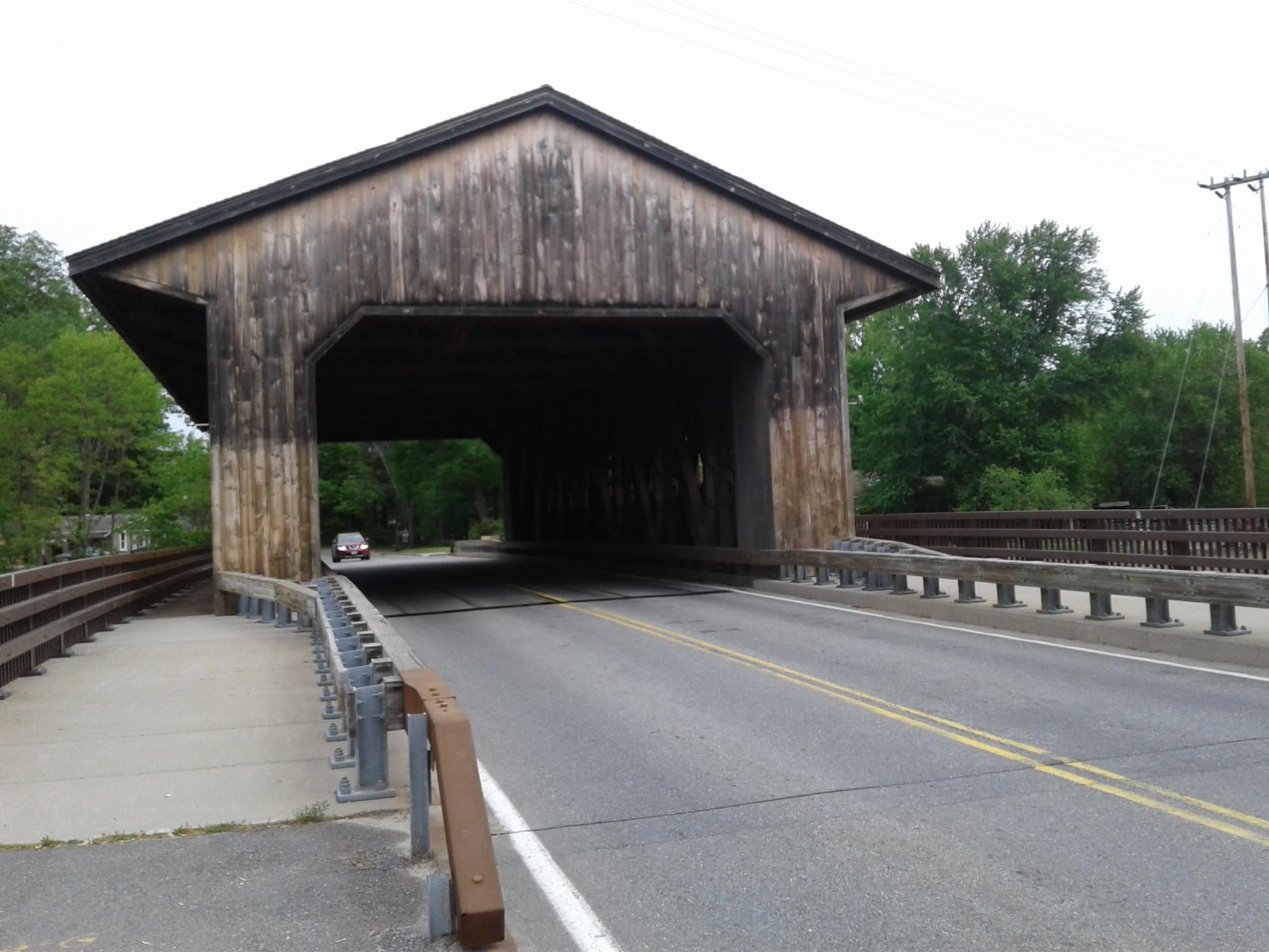 A rustic wooden covered bridge over a road, with a car approaching. - Pepperell Covered Bridge – June's Wood loop from Groton