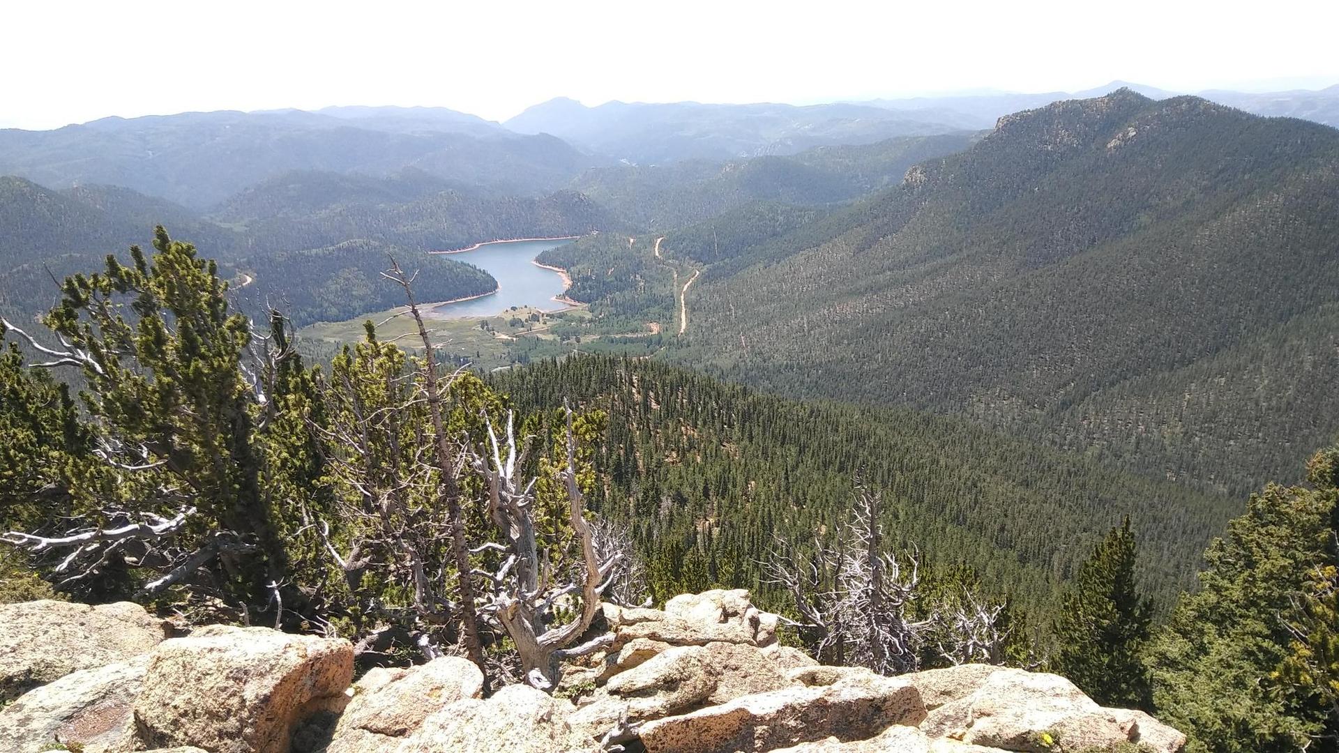 Panoramic view of a forested valley with a winding lake, from a rocky peak.