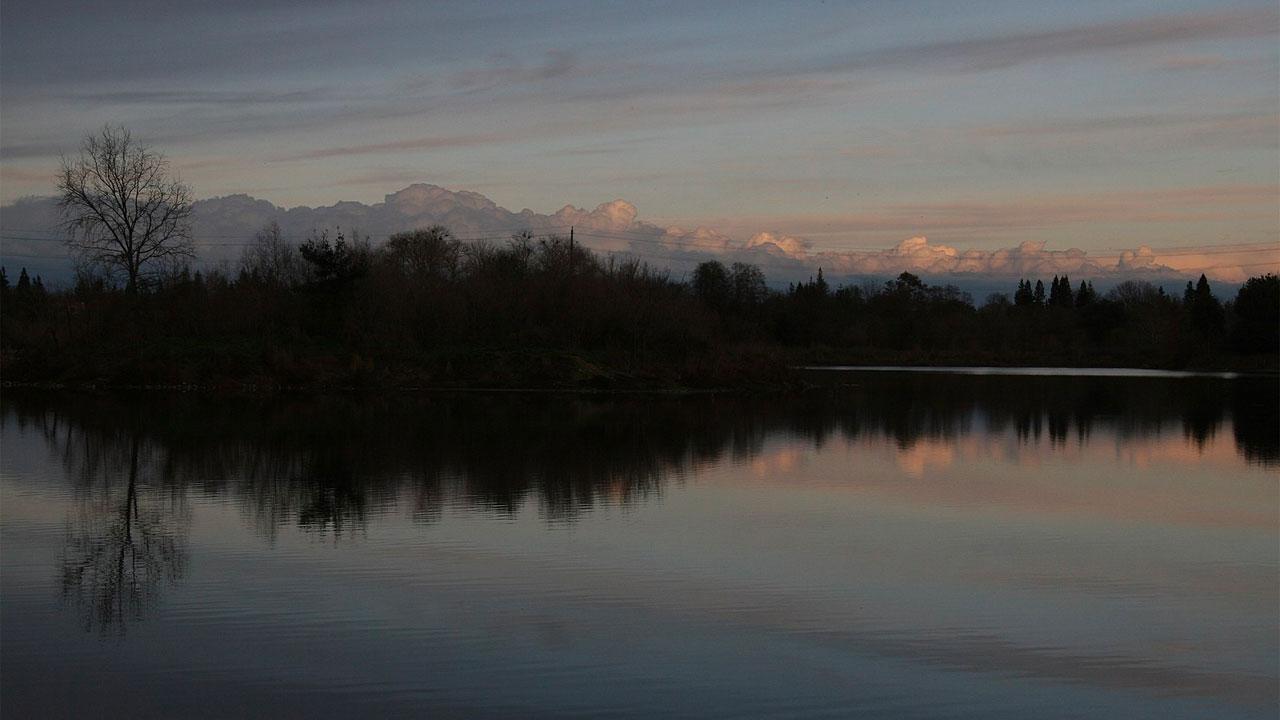 Calm lake reflecting dark trees and distant mountains under a twilight sky.