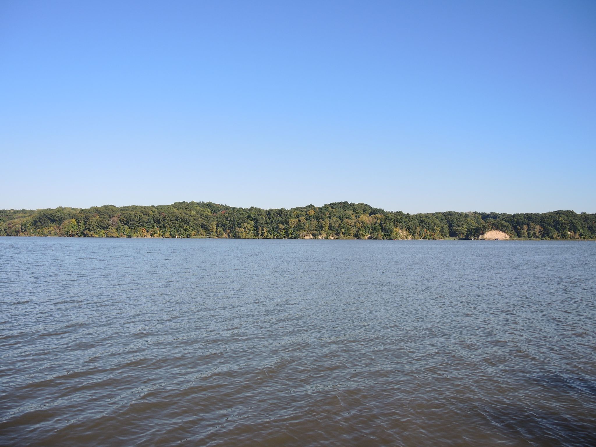 Wide shot of calm water with a distant green forested shoreline under clear sky. - Irondequoit Bay loop