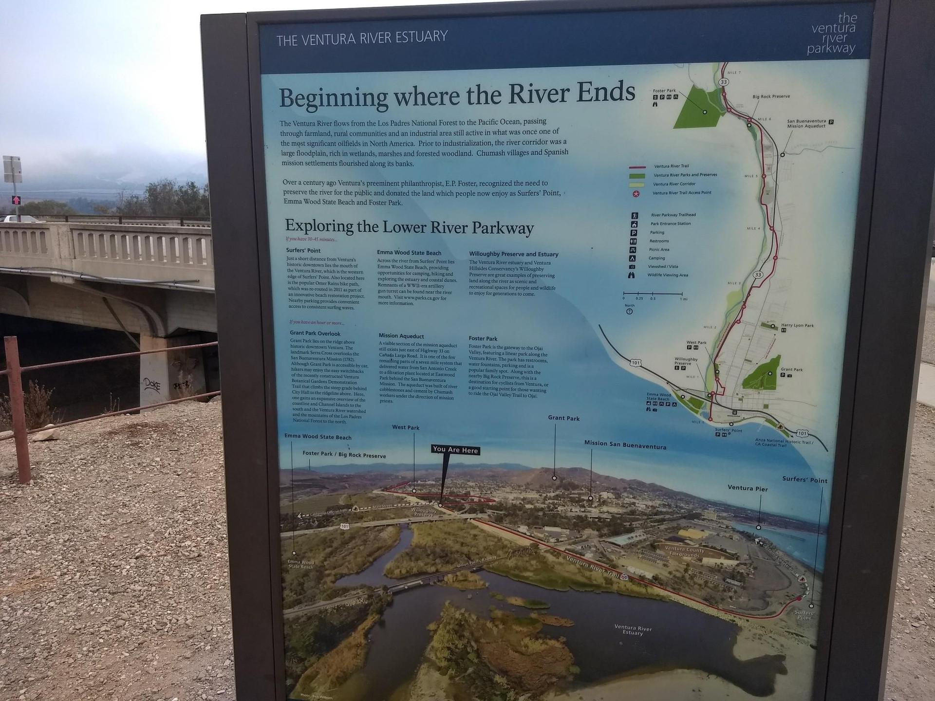 Information sign with maps and text, a bridge in the background.
