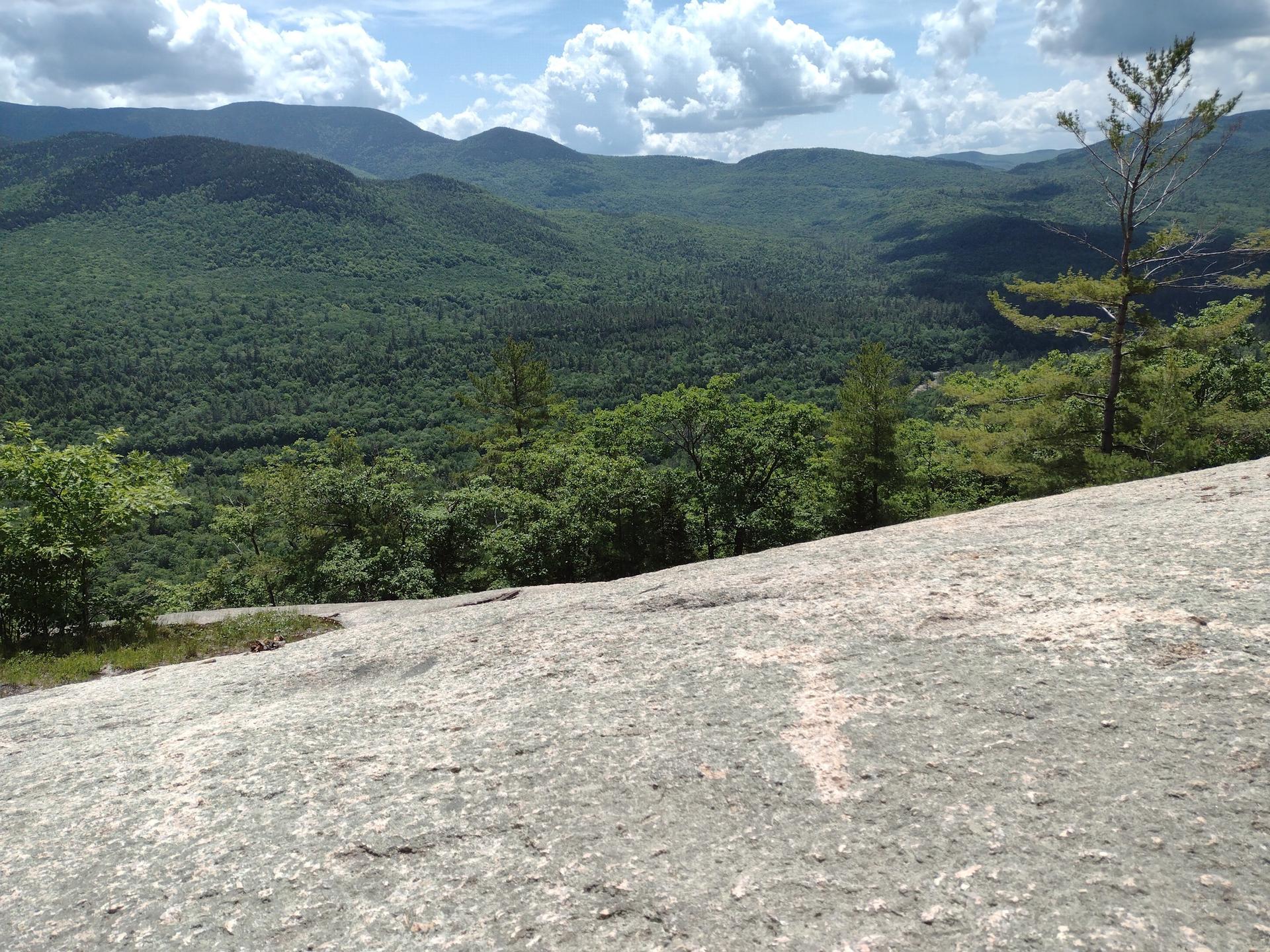 Rocky outcrop overlooking lush green forested mountains under a partly cloudy sky.