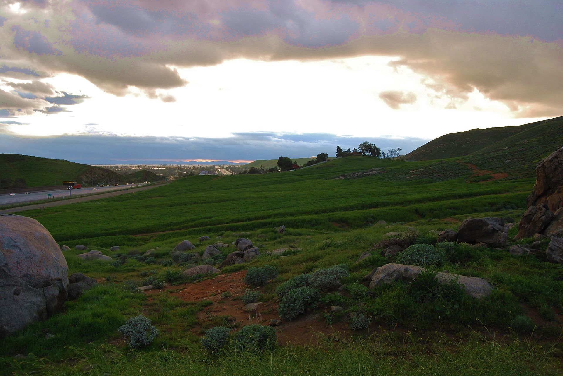 Lush green hills and large boulders under a dramatic, cloudy sky with a distant highway. - Oak Quarry Golf Club to Jurupa Mountains Discovery Center