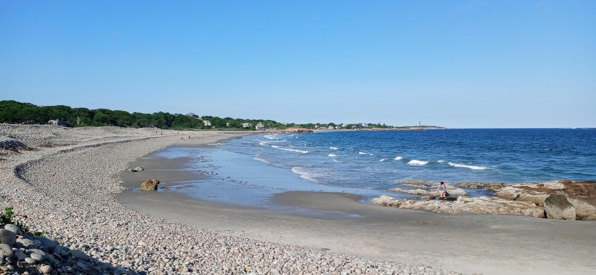 Wide pebbly and sandy beach with ocean waves under a clear blue sky.