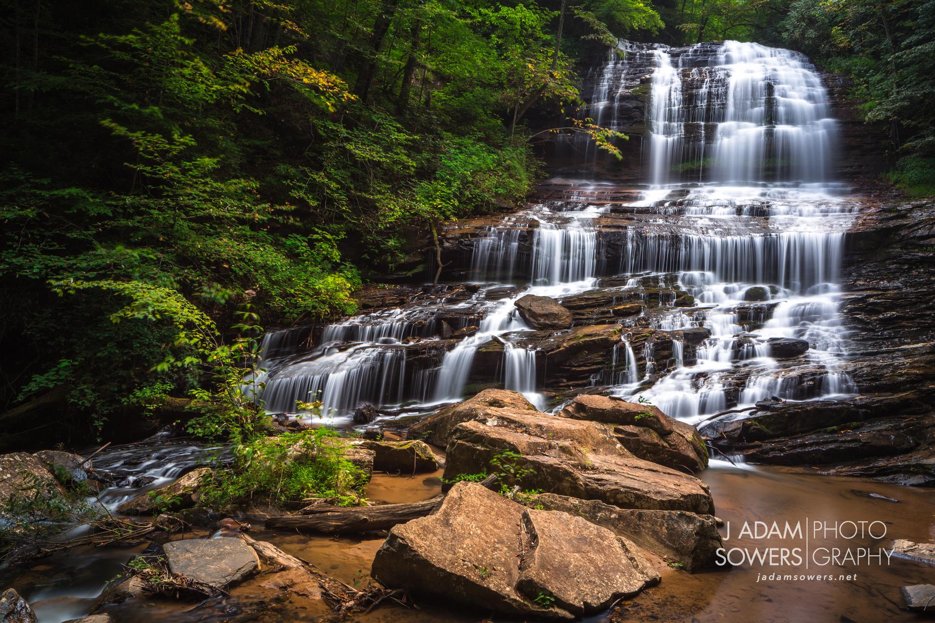 A multi-tiered waterfall cascades over rocks in a lush green forest.