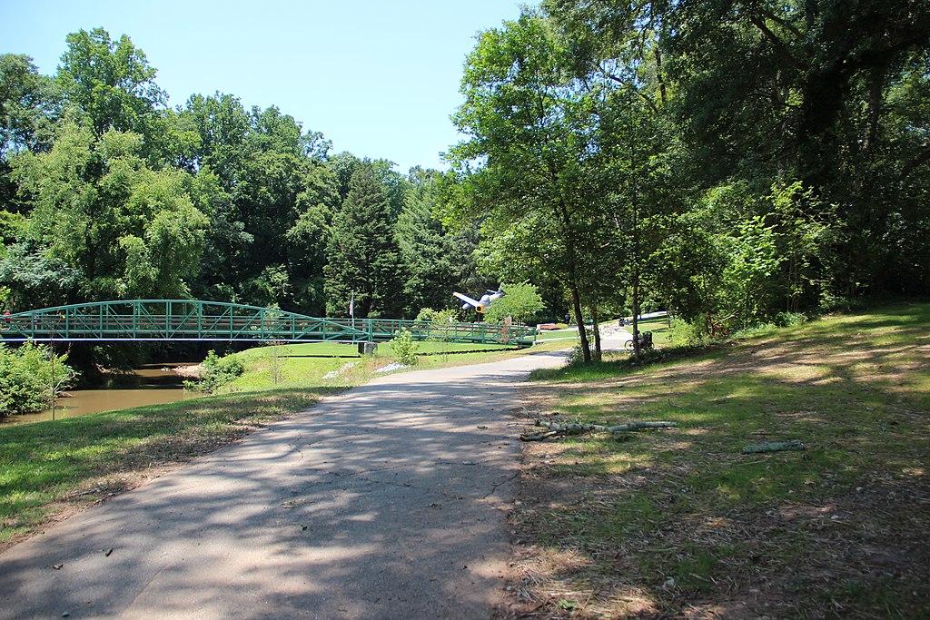 Green bridge over a river, dirt path, surrounded by lush trees in bright daylight.
