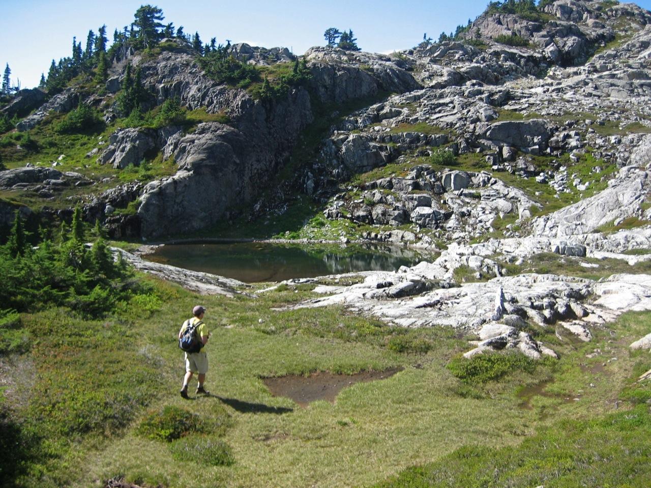 A hiker walks past a small lake in a sunny, rocky mountain landscape.