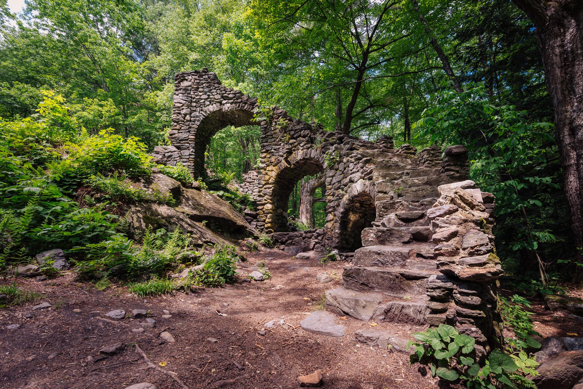 Stone ruins with arches and stairs in a lush green forest with dappled light.