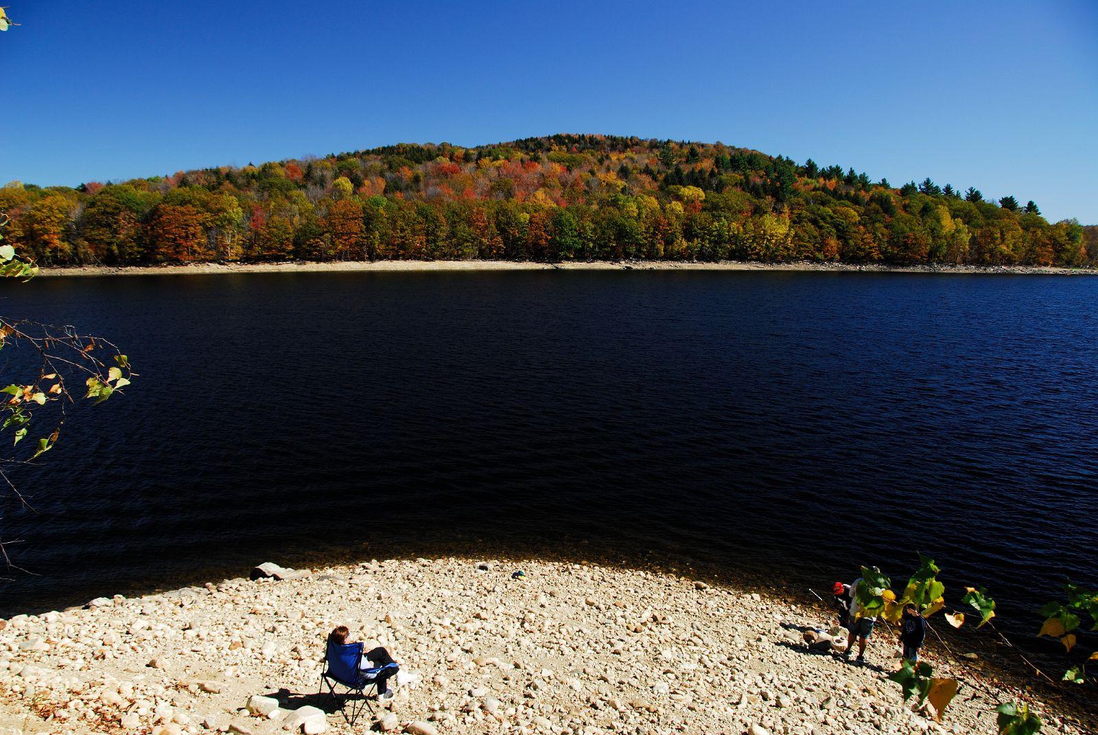 People on a rocky shore beside a dark lake and colorful autumn forest.