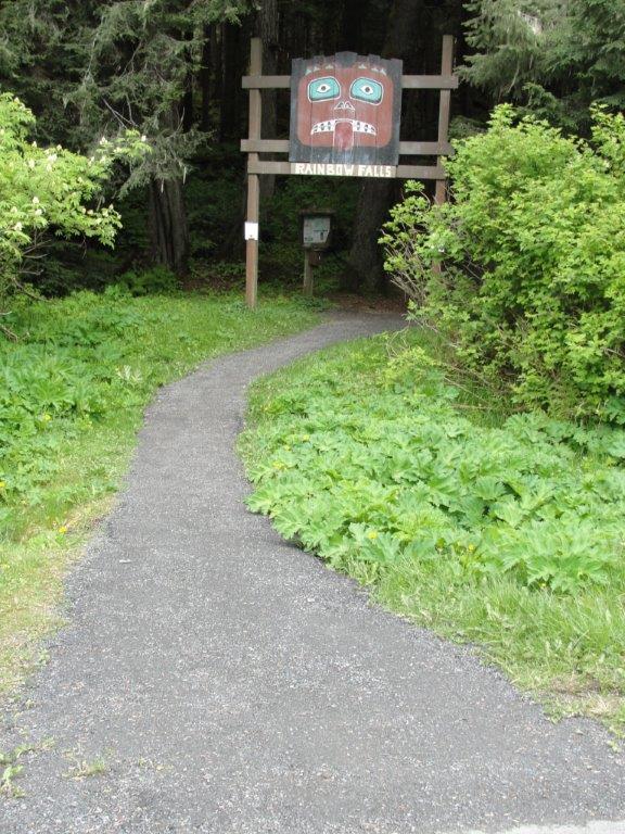 Gravel path leading to a wooden sign with a carved face amidst green foliage.