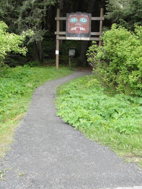 Gravel path leading to a wooden sign with a carved face amidst green foliage. - Rainbow Falls Trail