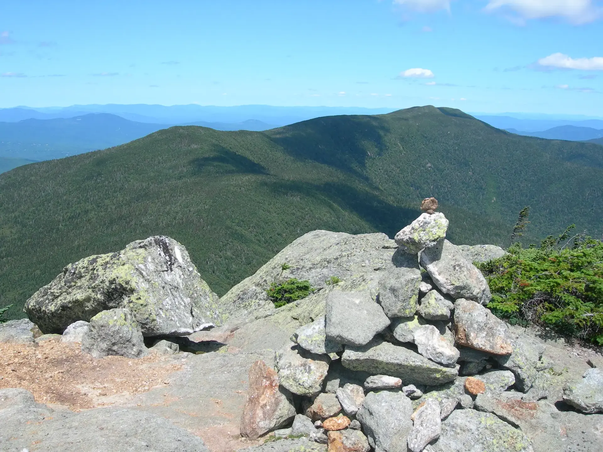 Rock cairn on a sunny, rocky mountain peak overlooking green valleys.