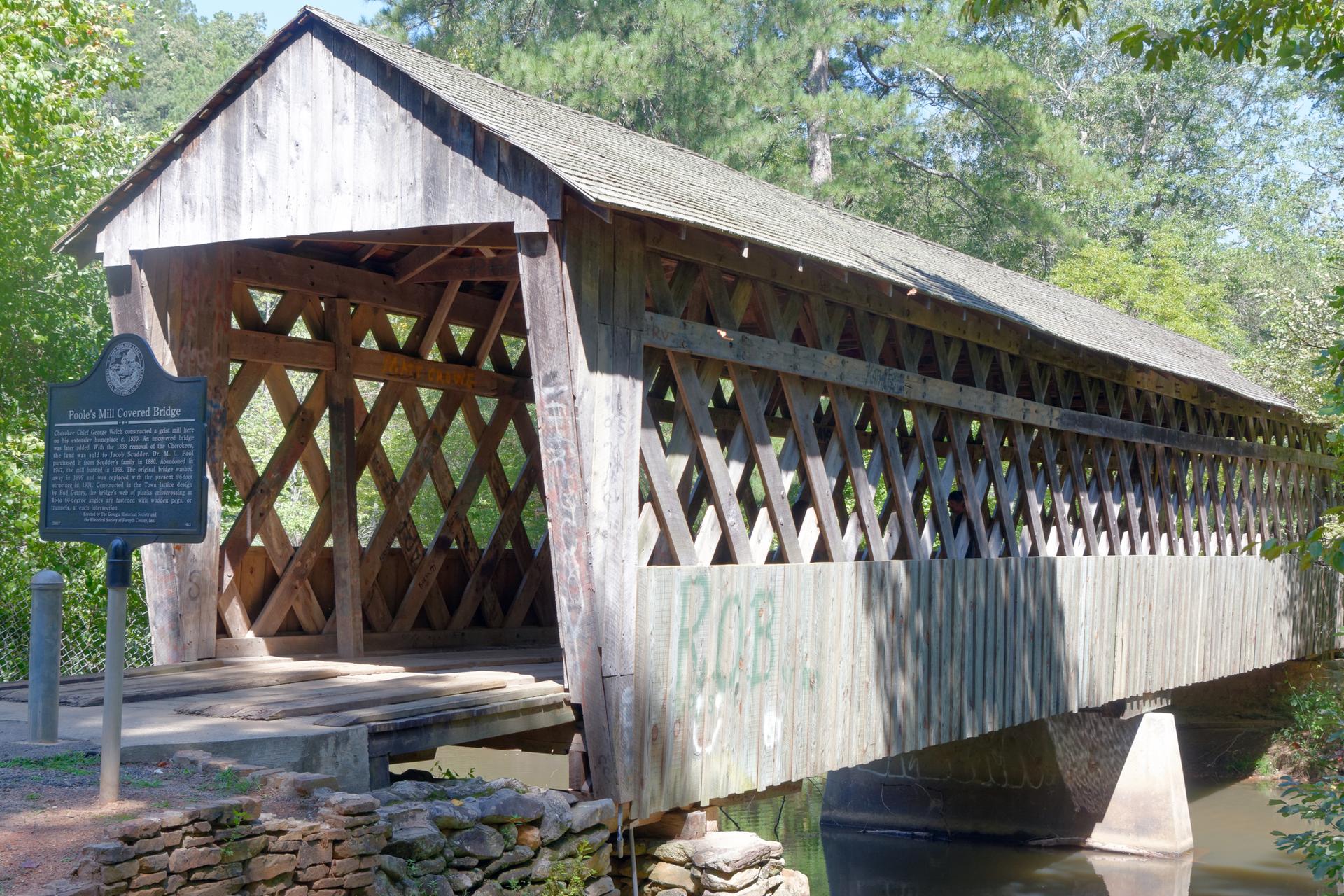 Rustic wooden covered bridge over water, with a historical marker, under bright sun.