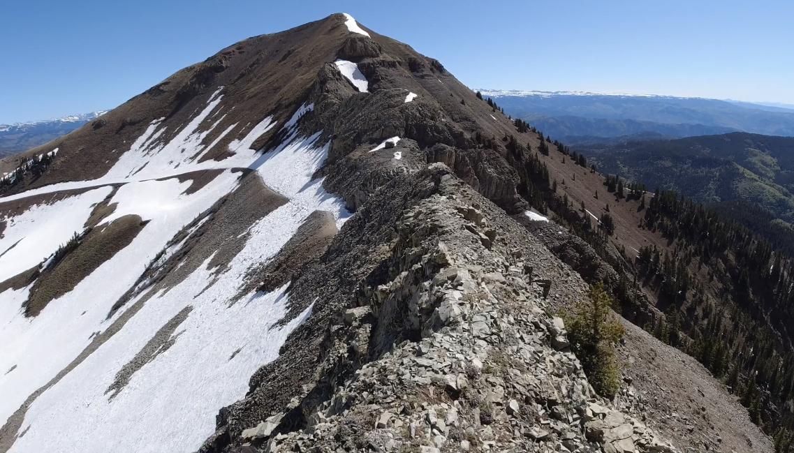 Rocky mountain ridge leading to a snow-dusted peak under a clear blue sky.
