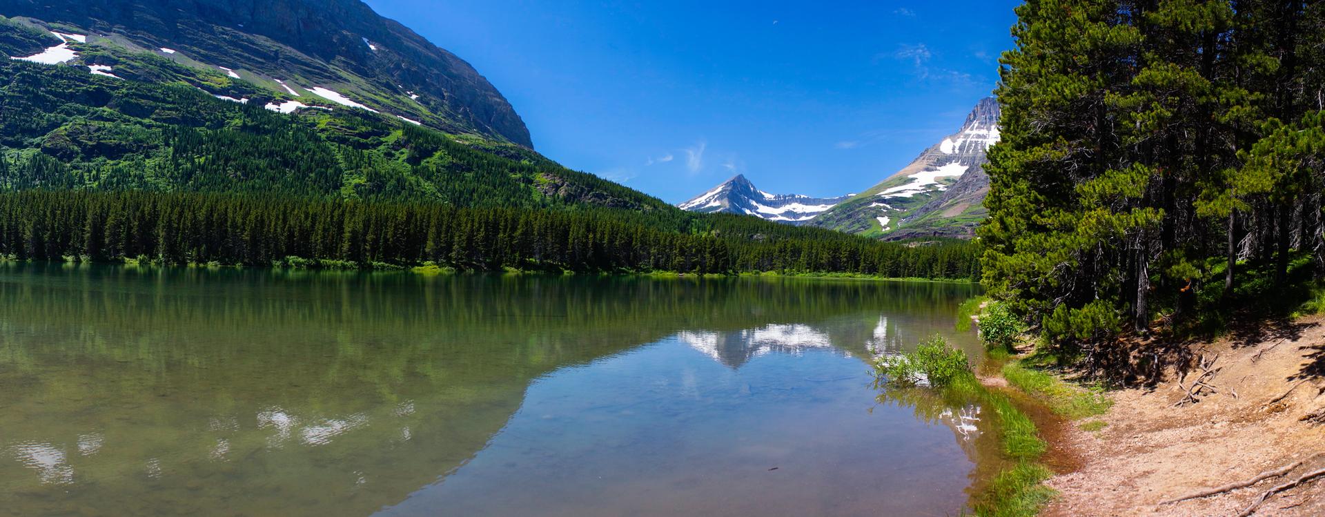 A serene lake reflects snow-capped mountains and a dense evergreen forest.