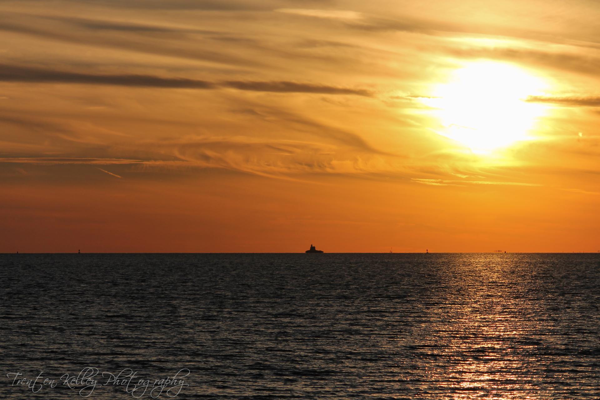 Vibrant orange and yellow sunset over calm water with a distant ship.