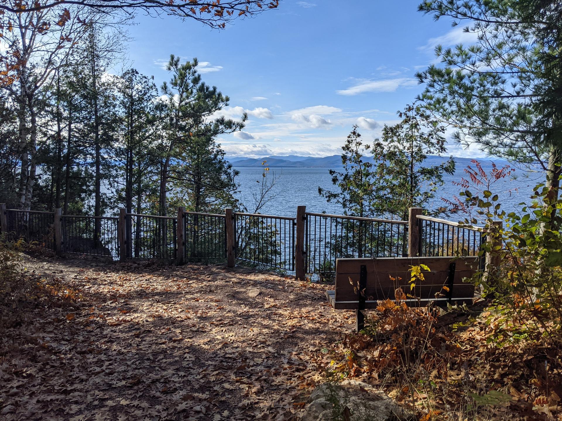 Forested overlook with a bench and fence, offering views of a large lake.