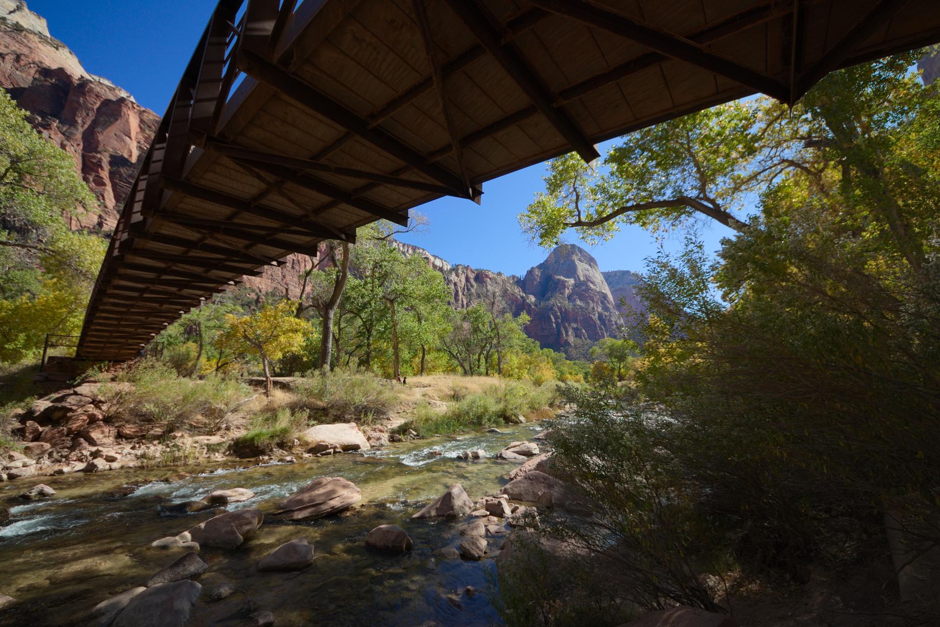 View from under a bridge over a rocky river, with mountains and trees.