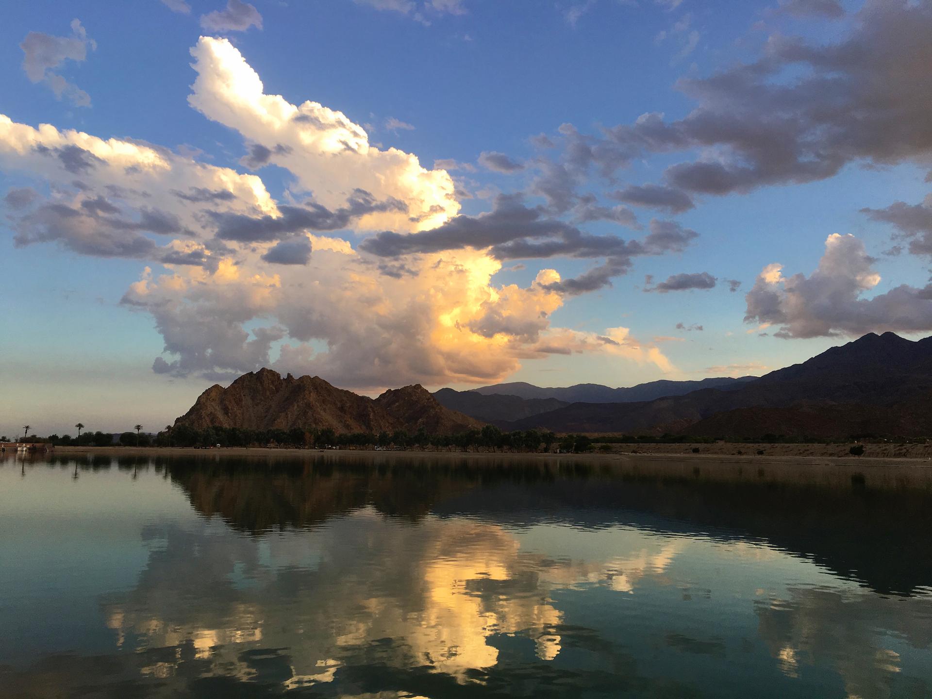 Calm lake reflecting golden clouds and mountains under a blue sky.