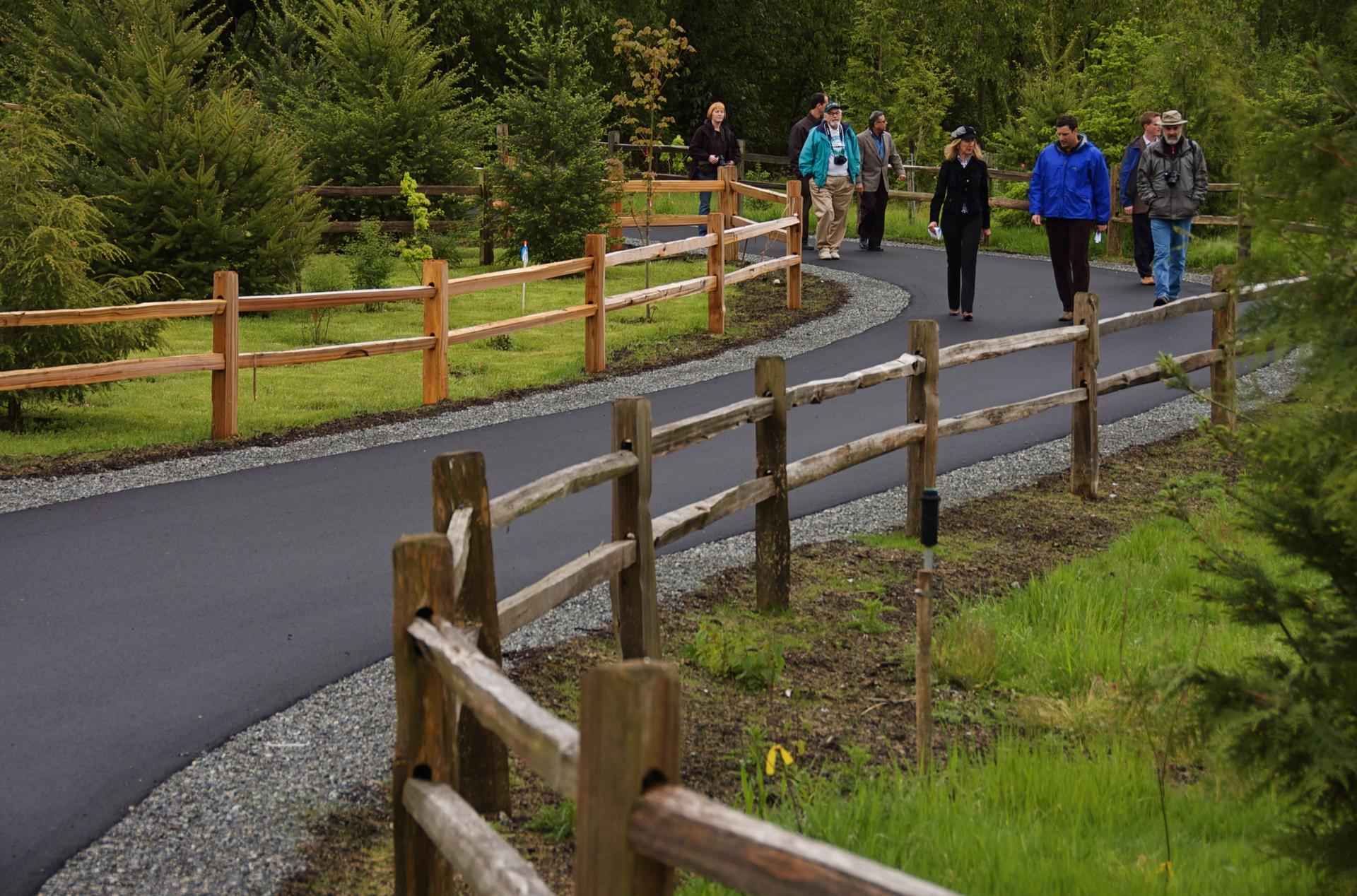 Group of people walking on a paved path through a green, fenced, wooded area.