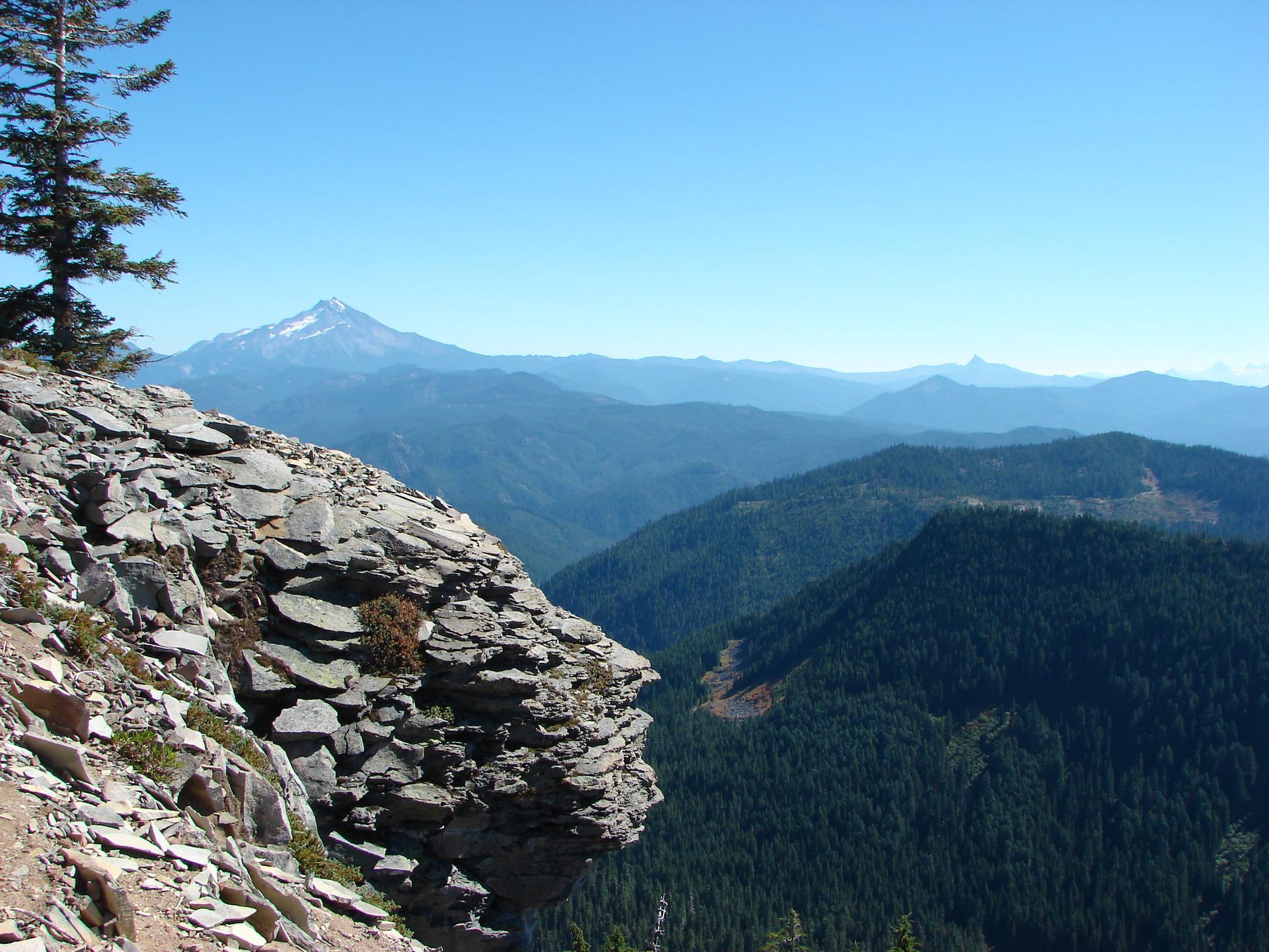 Rocky cliff edge overlooking vast forested valleys and distant mountains on a clear day.