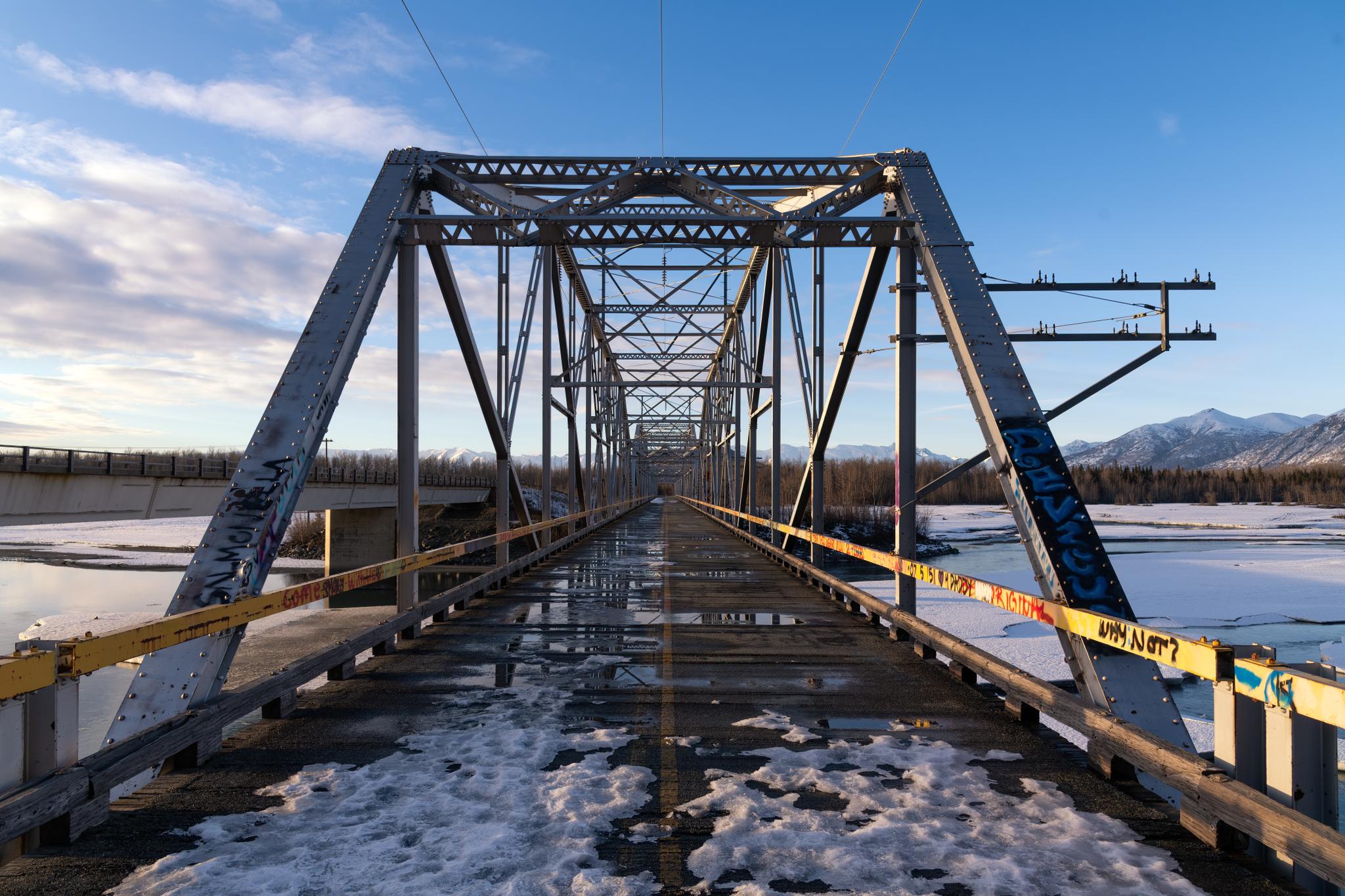 Long metal truss bridge over a snowy river under a blue sky.