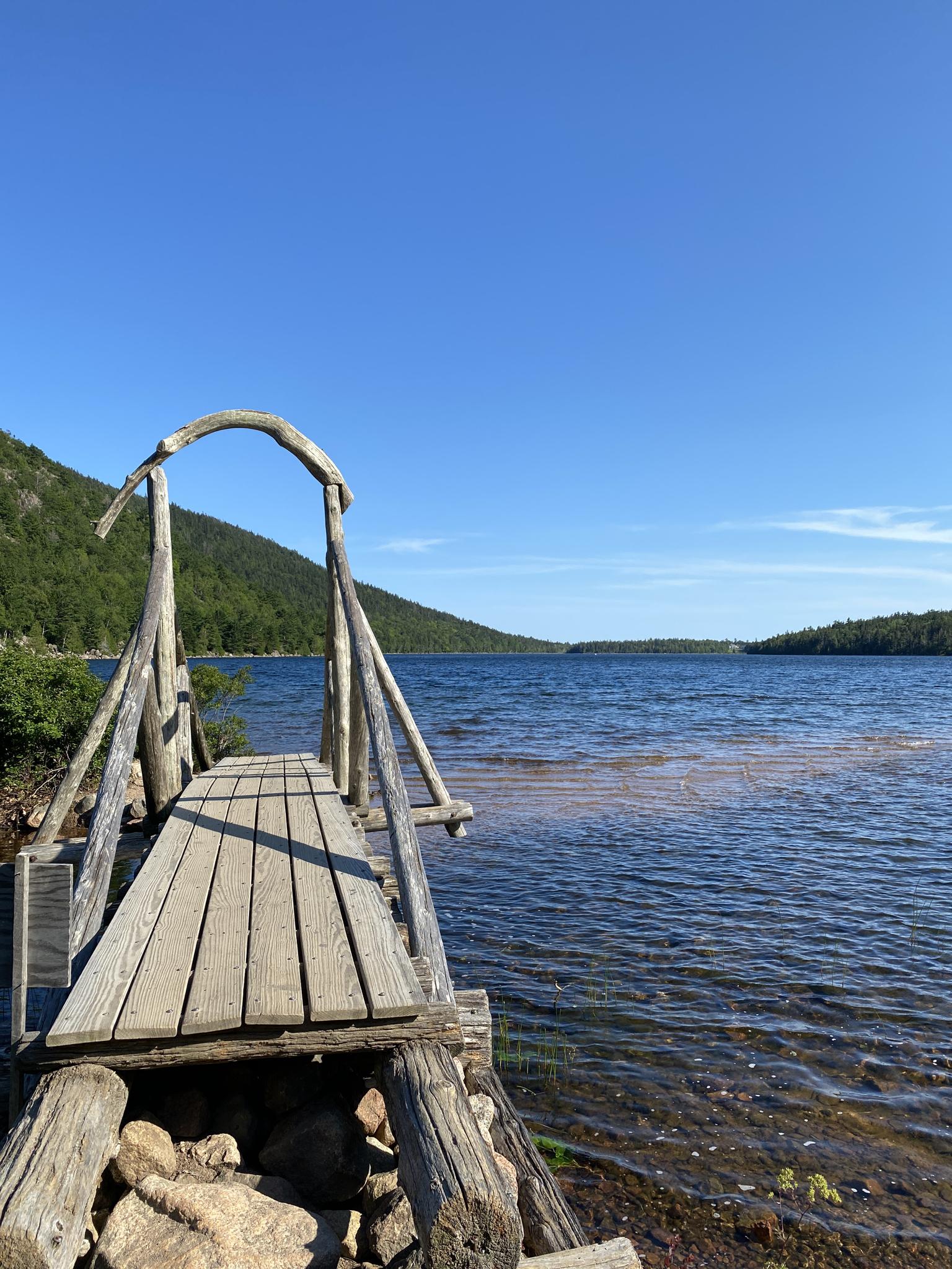 Wooden footbridge over clear lake water, surrounded by green forested hills.