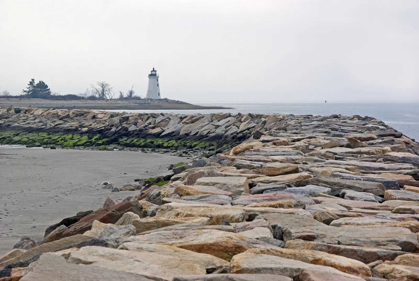 Stone jetty extending into calm water towards a distant lighthouse under an overcast sky.