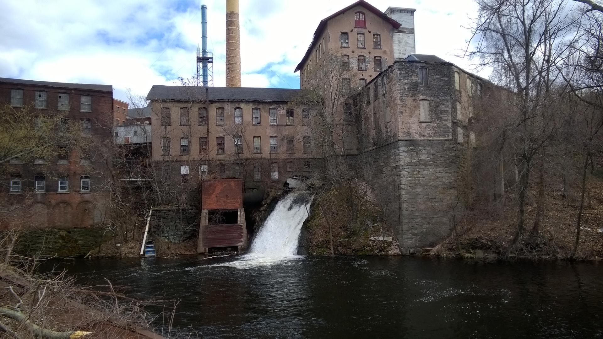 Old industrial buildings with a waterfall flowing into a river under an overcast sky.