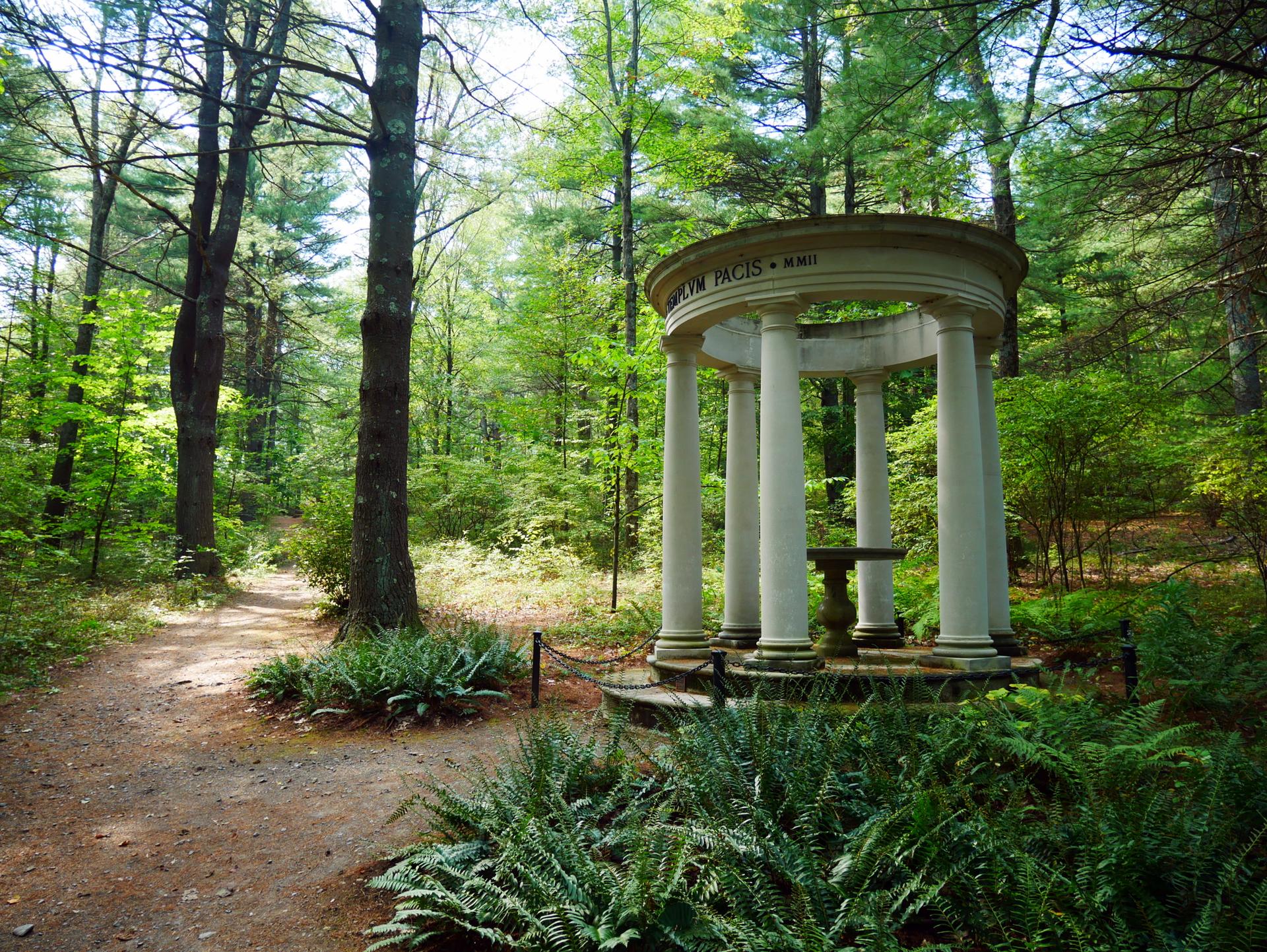 A white columned gazebo stands in a sunlit green forest with a dirt path.