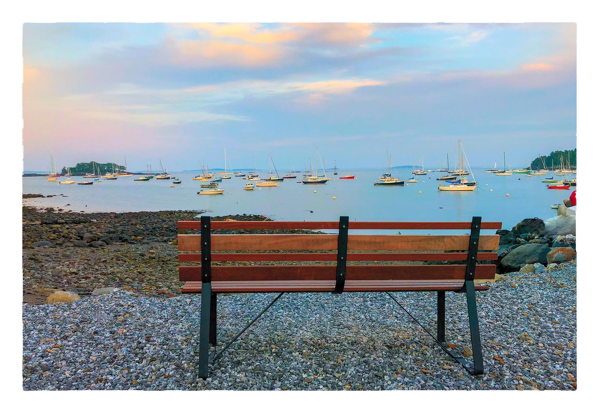Empty bench on a rocky shore overlooking boats at sunset.