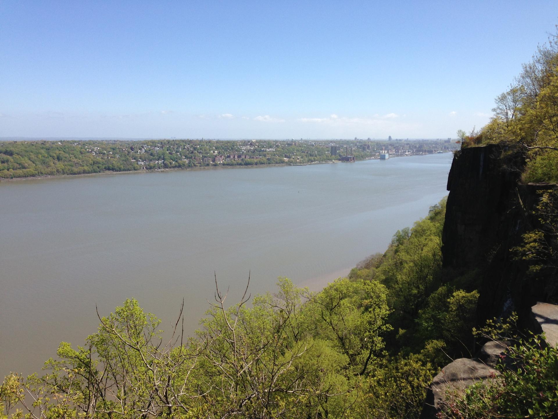Sunny view of a wide river, green trees, distant city, and rocky cliff.