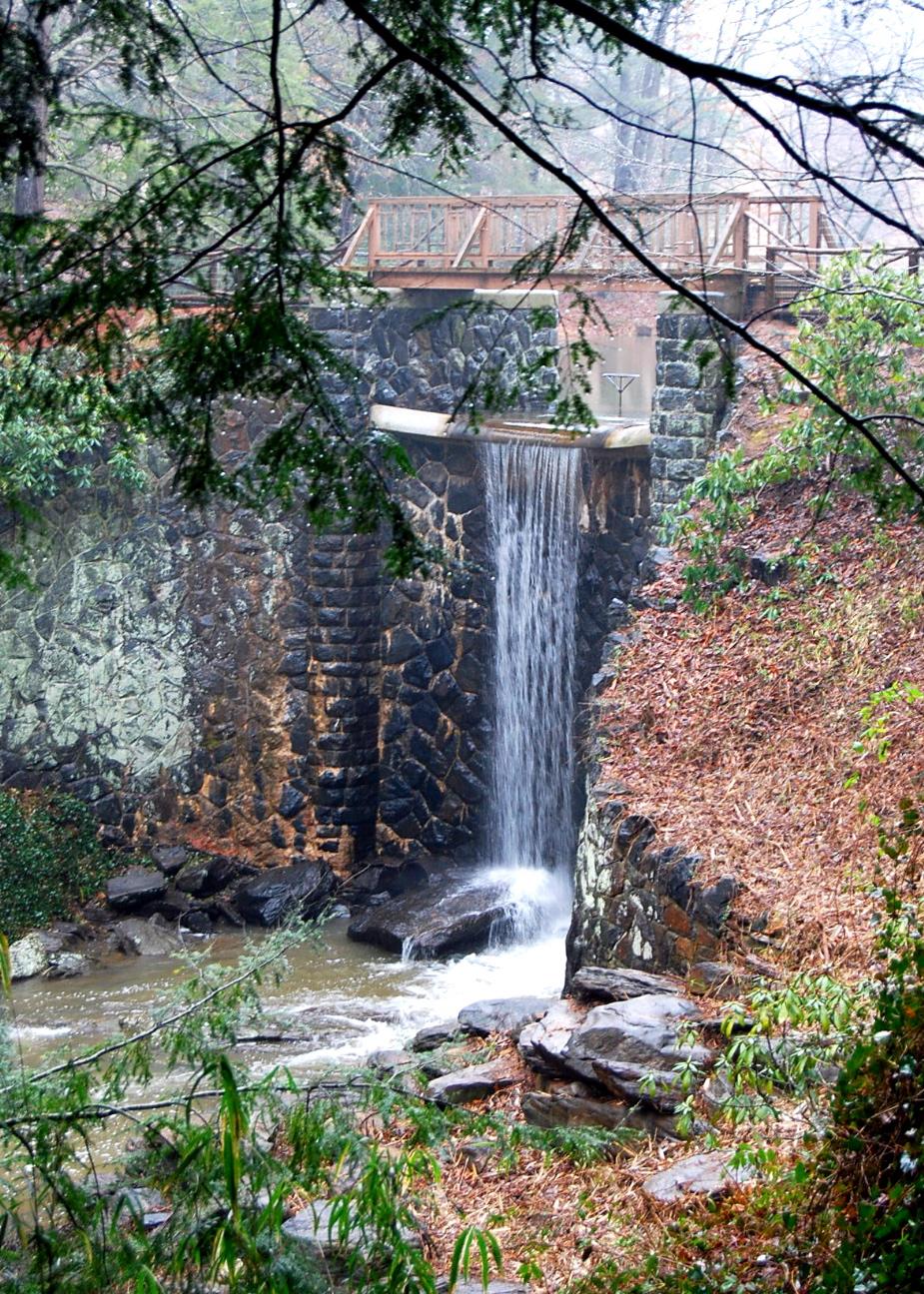 Waterfall cascading over a stone wall into a rocky stream, framed by trees.