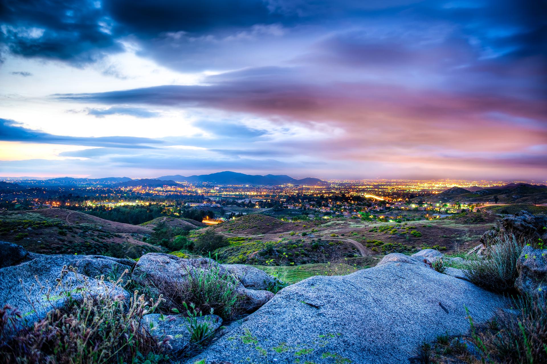 Rocky foreground overlooking a city at twilight with colorful, cloudy skies.