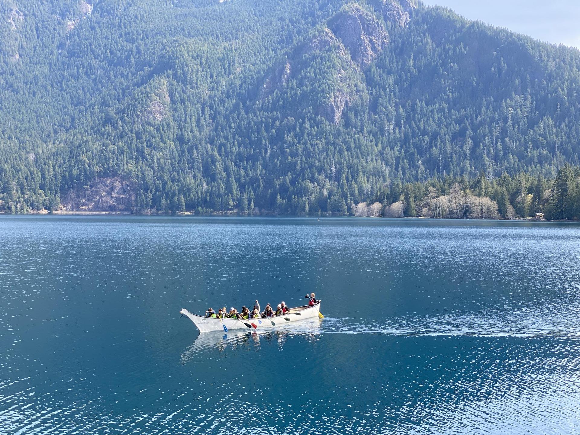 Long canoe with people paddling on a blue lake, surrounded by forested mountains.