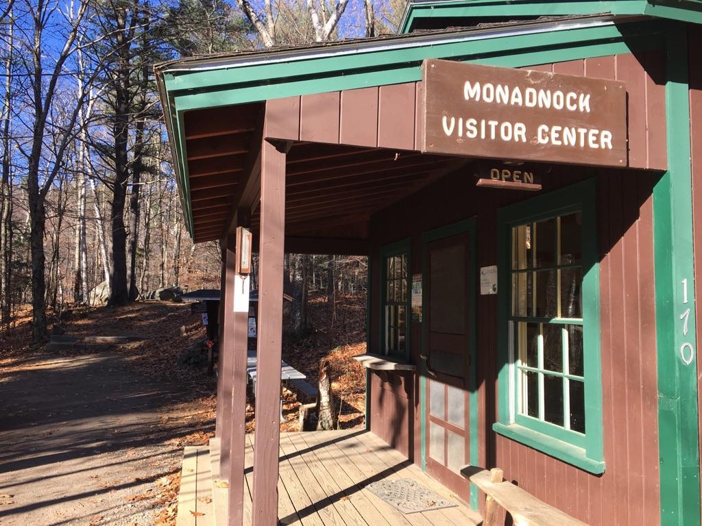 Brown wooden building with green trim, porch, and forest background on a sunny day.