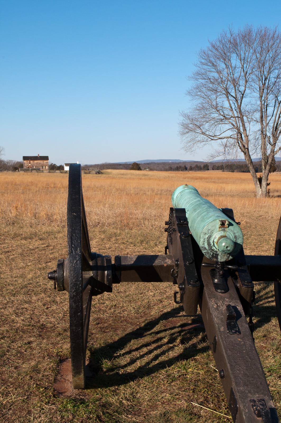 Cannon in a dry grassy field with a distant house under a clear sky.