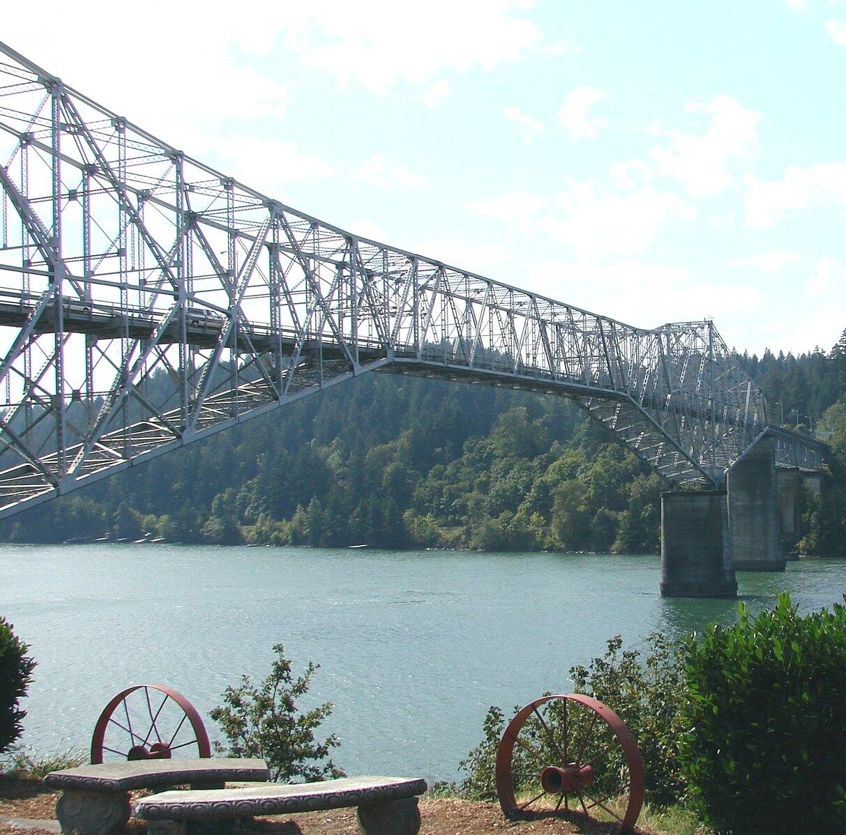 Large steel truss bridge spans wide river with forested hills under bright sky.