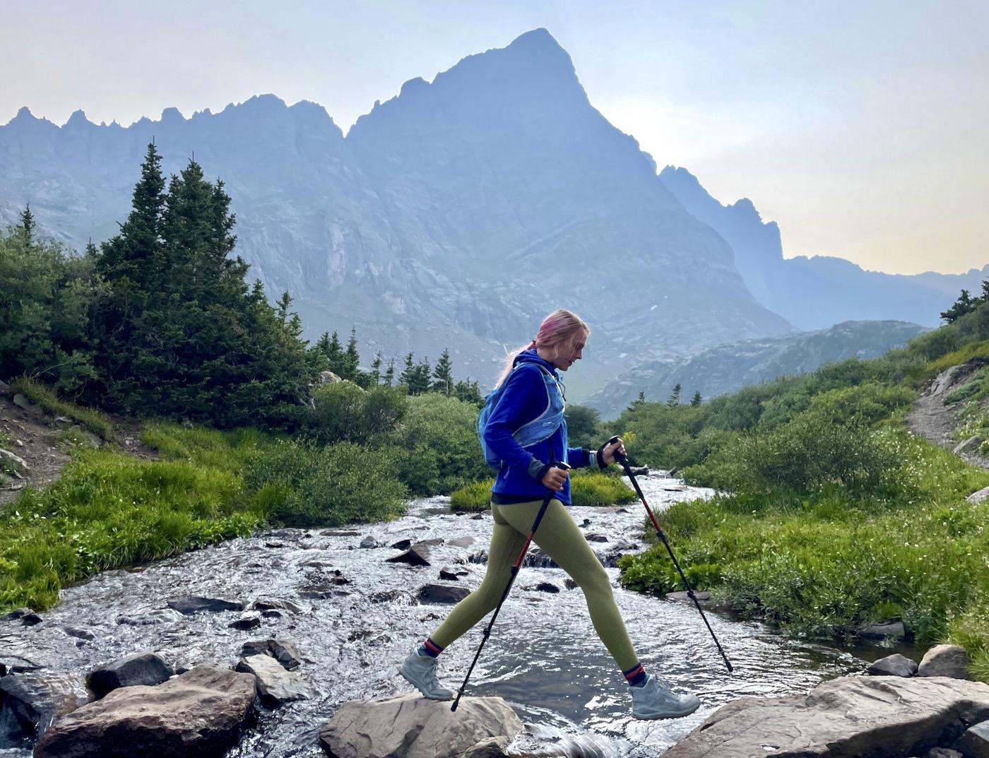 Hiker with poles carefully crosses a rocky stream in a vast mountain landscape.